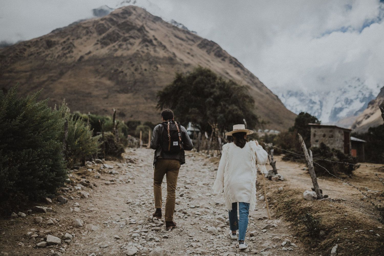 Two travelers walking along dirt path through mountainous terrain.