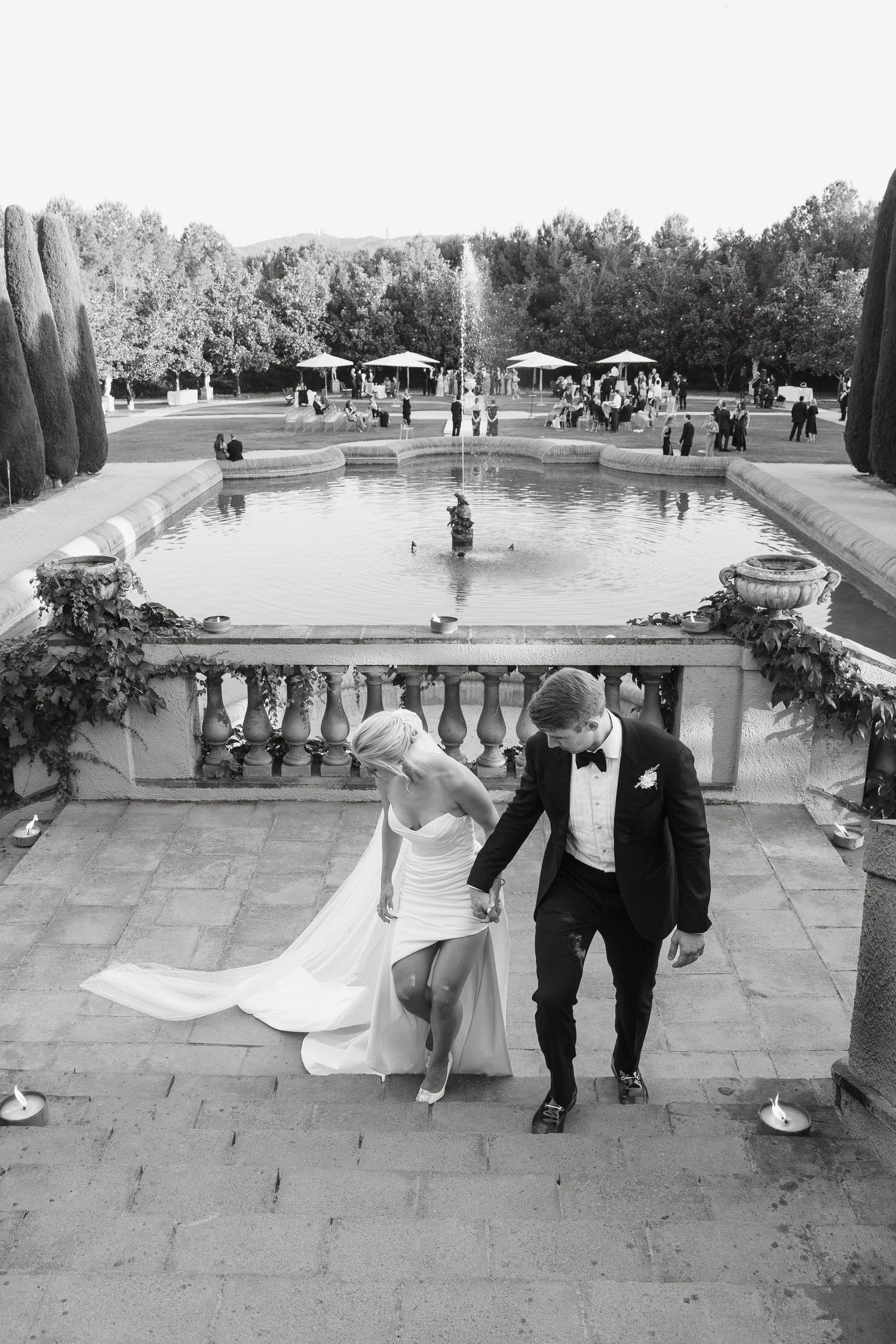 Newlyweds descending stone steps with a fountain and garden party in the background.