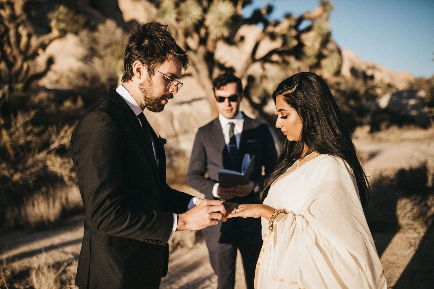 A romantic desert wedding ceremony takes place among rocky formations at sunset with warm golden lighting.