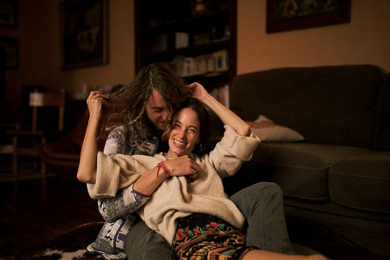 Playful couple lounging and laughing together on a dark sofa in a warm living room setting.