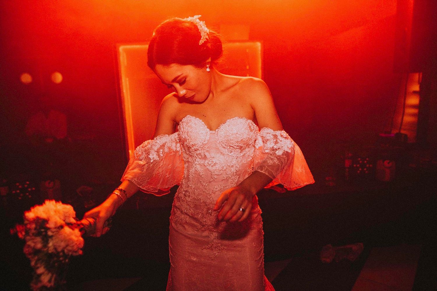 Bride in off-shoulder lace gown illuminated by dramatic red lighting.