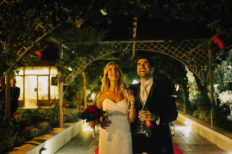 Evening garden wedding scene with couple beneath lit archway and string lights.