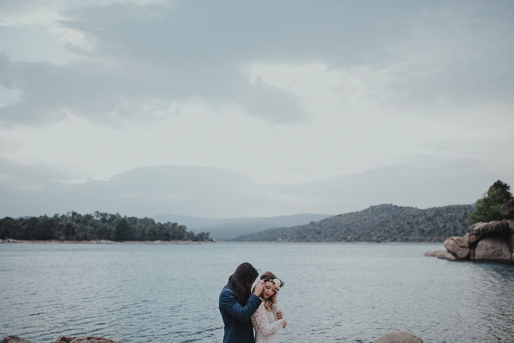 Two people share a romantic moment on rocky shores overlooking a serene mountain lake.