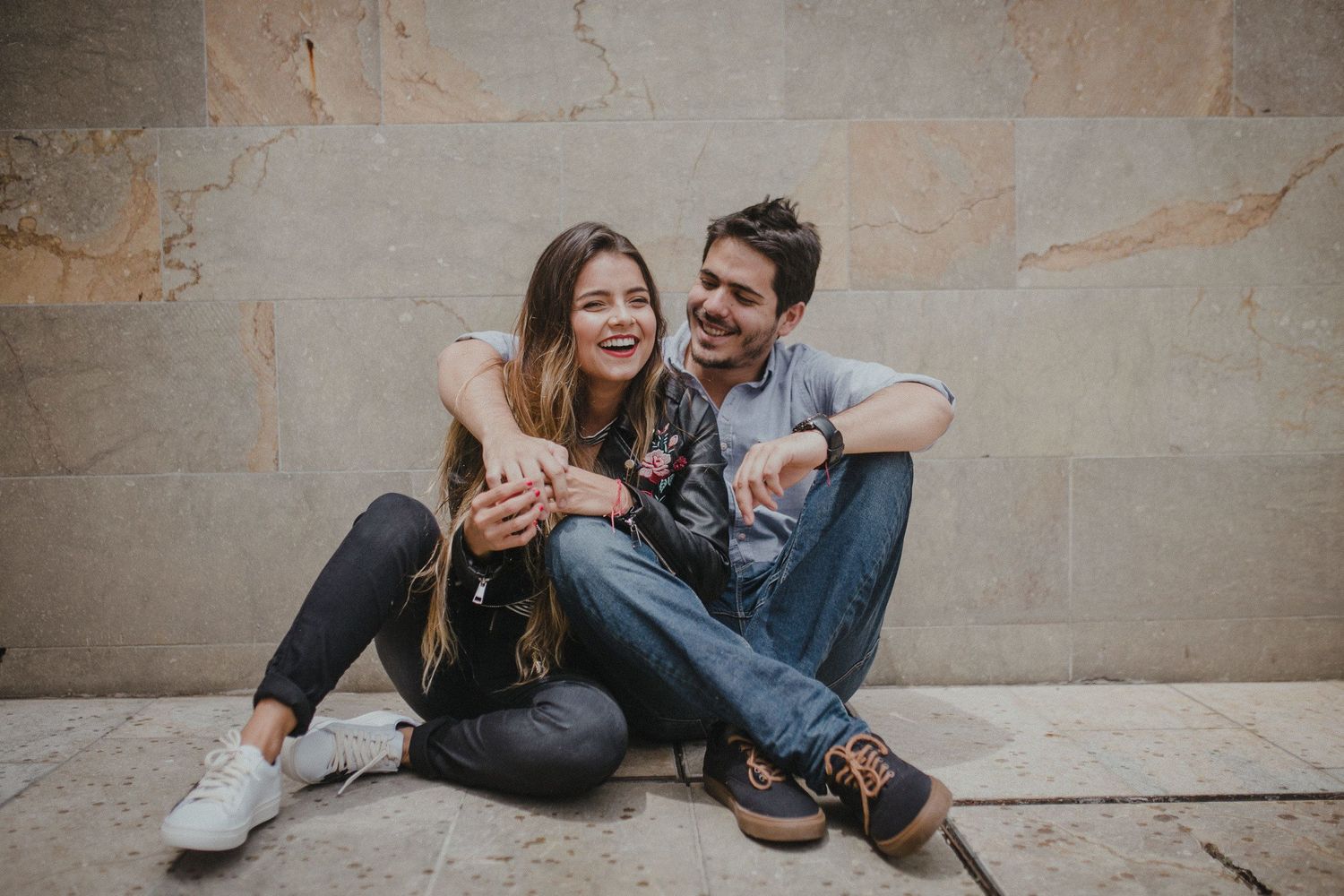 A casual couple sits against a wall laughing while looking at their phone together.