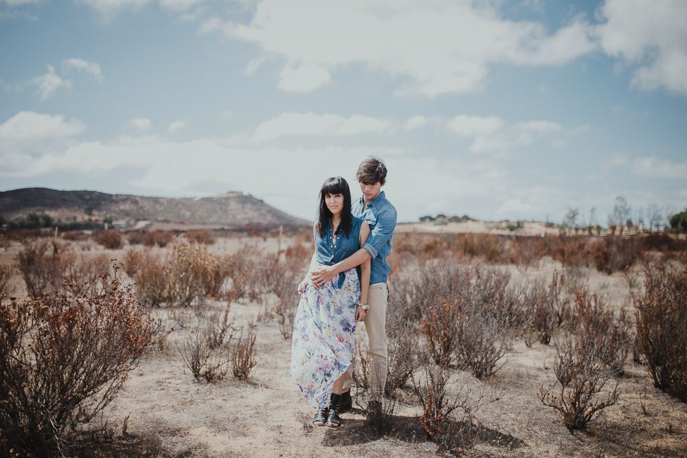 Two people embrace while standing among desert brush with mountains visible in the distance.