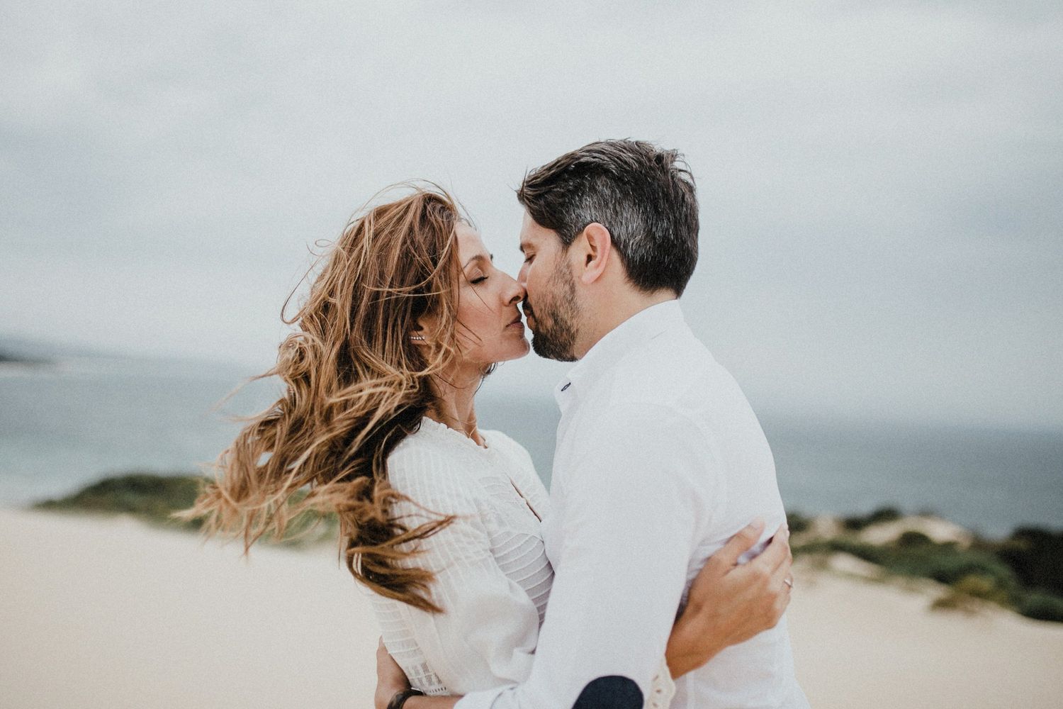 Romantic beach portrait with flowing hair and white outfits against a misty ocean background.