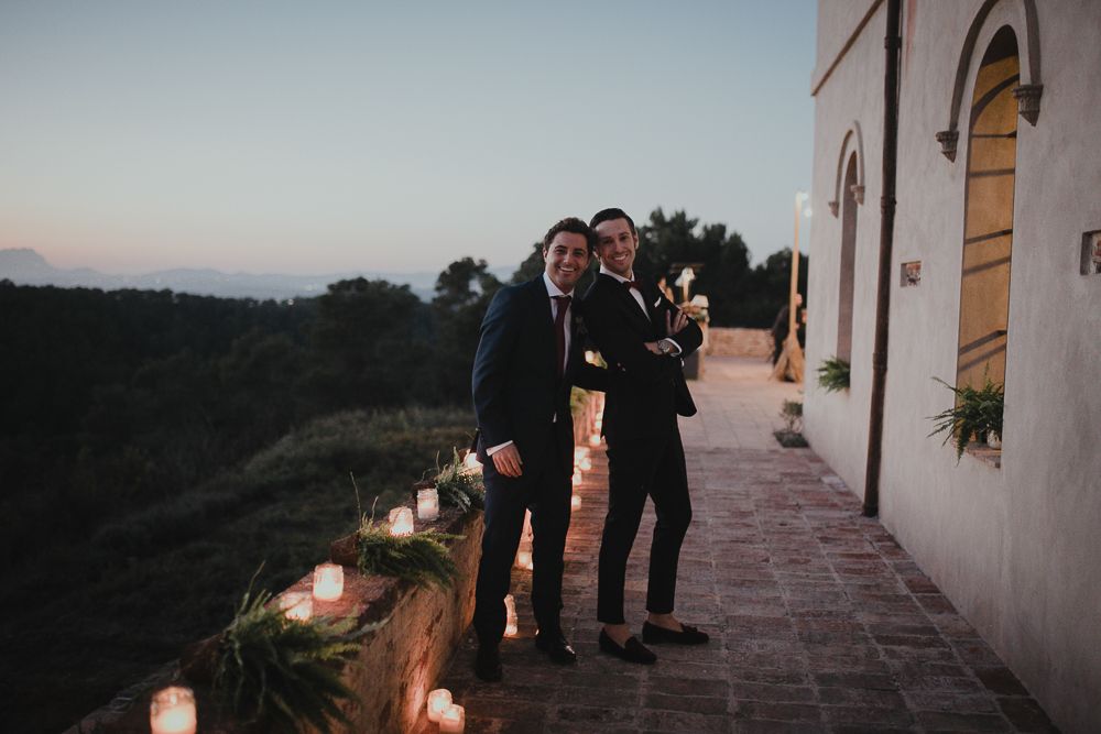 Two figures stand on a candlelit outdoor terrace at dusk next to an elegant white building with arched windows.