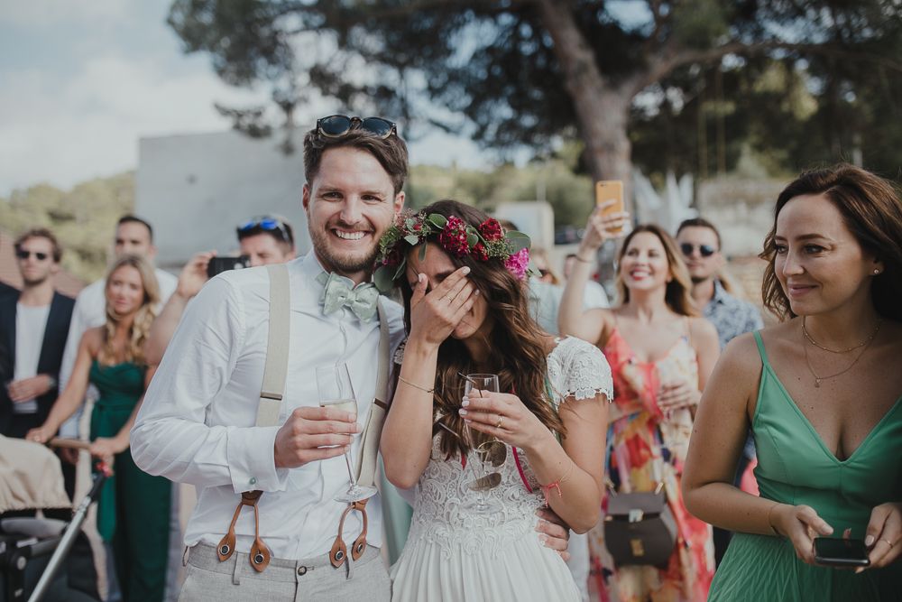 Wedding celebration outdoors with guests enjoying drinks and laughter in a sunny garden setting.