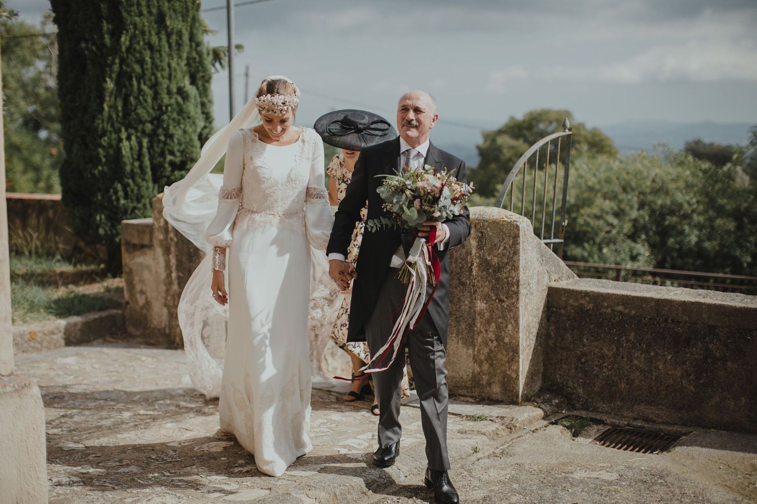 Elegant wedding couple walking along a stone pathway with cypress trees in background.