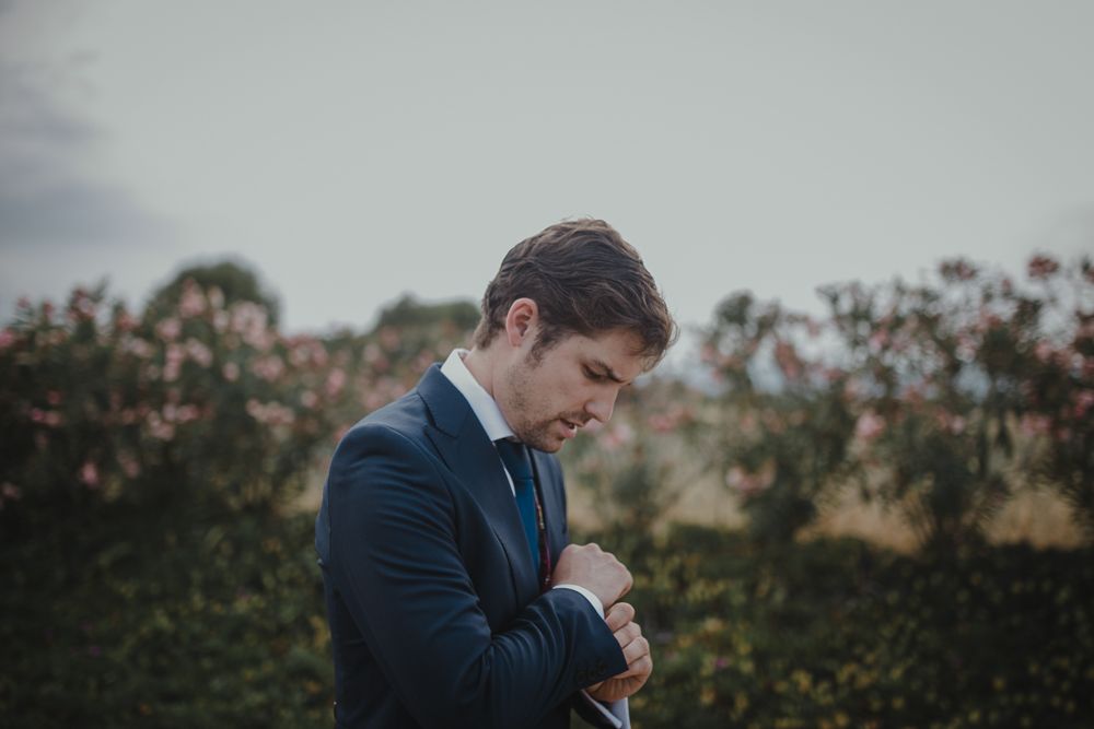 Person adjusting sleeve cuff in navy suit against rural background.