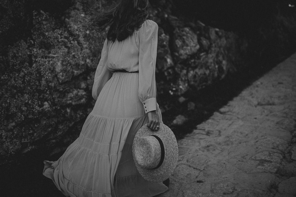 A flowing white dress and straw hat silhouetted against rocky terrain in black and white.