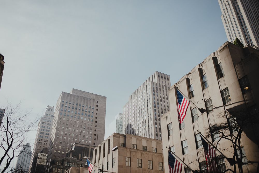 Low angle view of New York City buildings with American flags against blue sky.
