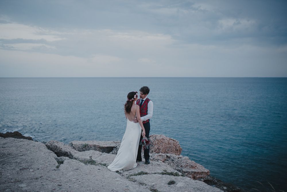 Wedding couple embracing on rocky oceanside cliff during overcast day.