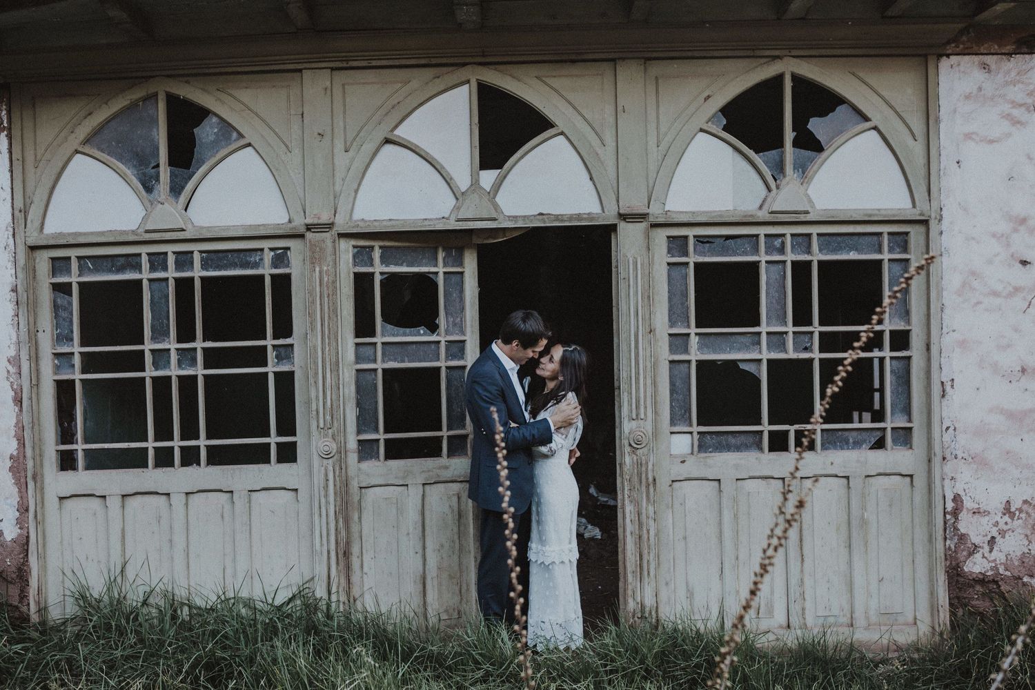 A romantic couple pose in front of vintage arched windows in an atmospheric outdoor setting.