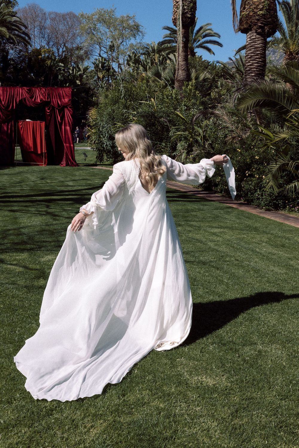 A person twirls in a flowing white dress on a manicured lawn with palm trees and red drapery.