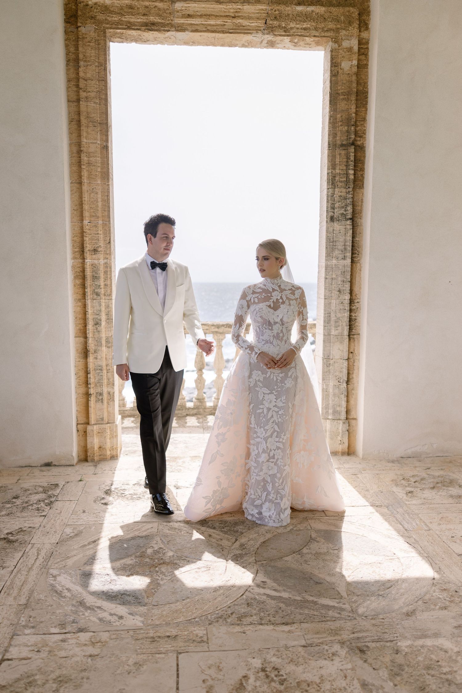 Elegant bride in lace long-sleeve gown and groom in white tuxedo jacket stand in stone archway with ocean view.