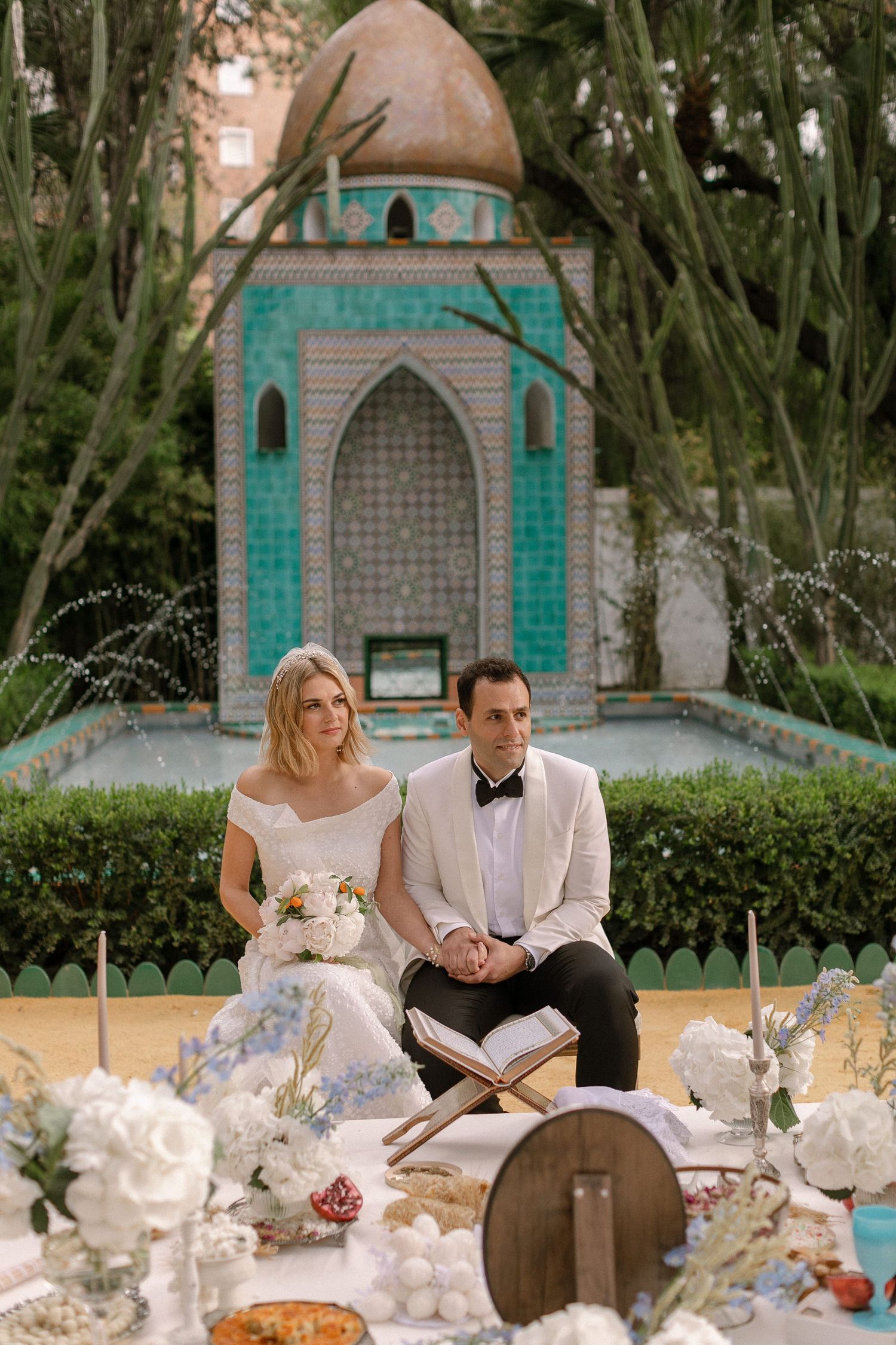 Bride and groom seated at decorated wedding table before ornate turquoise Moroccan fountain in lush garden setting.
