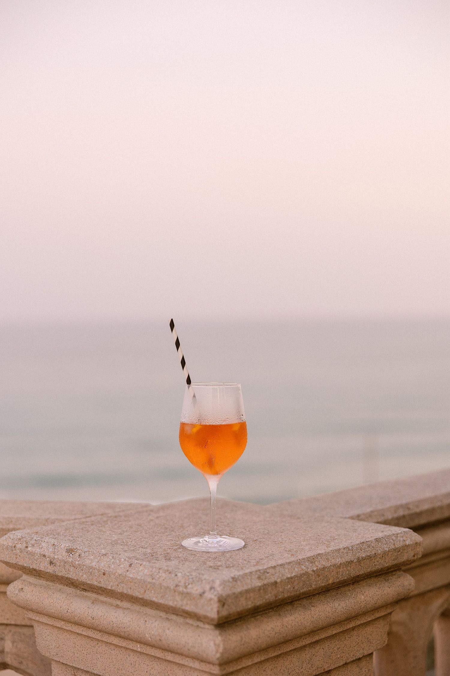 Orange cocktail in a wine glass with a striped straw on a stone balustrade overlooking a calm sea at dusk.