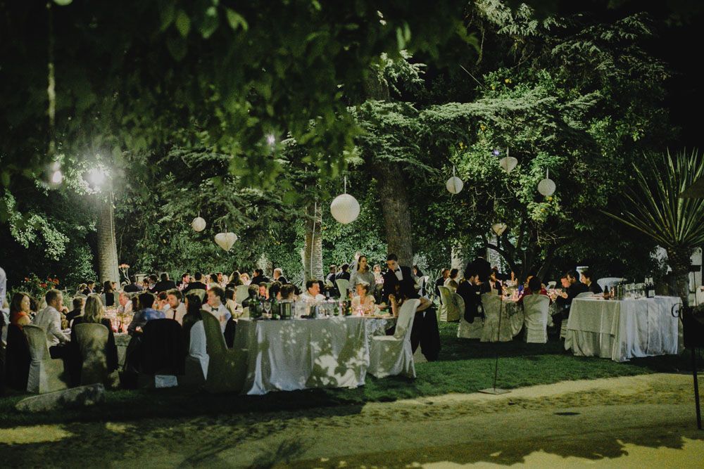 An elegant outdoor wedding reception at night with tables and hanging lanterns under lush greenery.