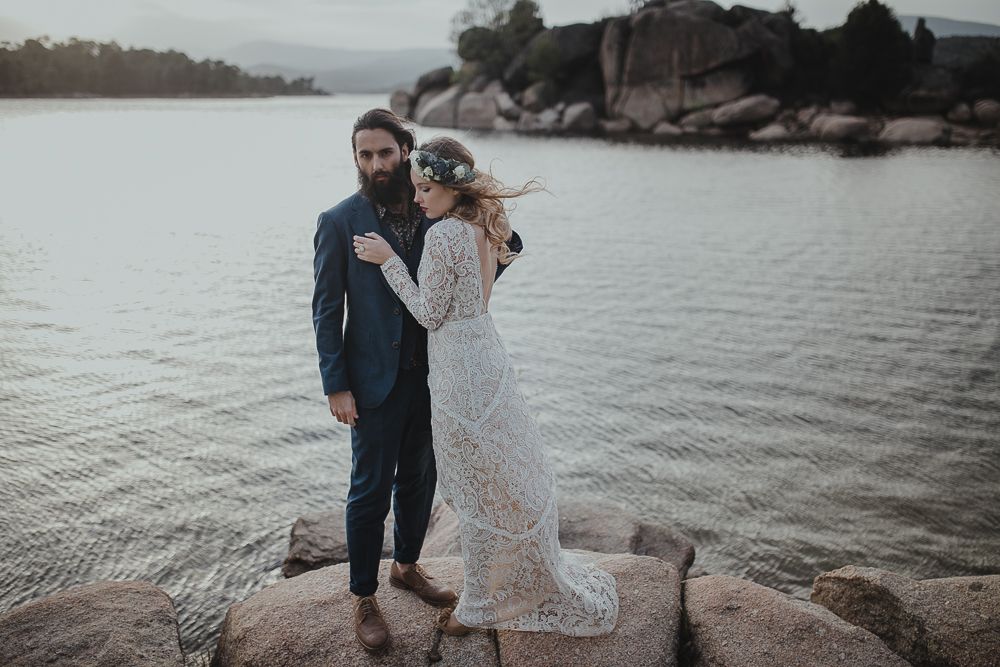 Couple stands on rocky outcrop with lake and mountains creating dramatic backdrop.