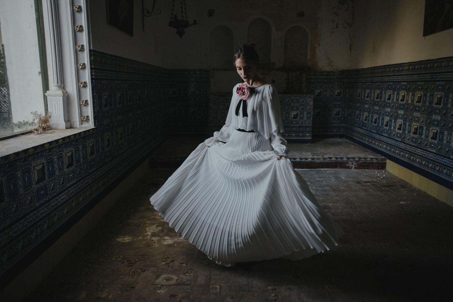A bride in a flowing pleated white gown twirls in a historic building with ornate dark walls.