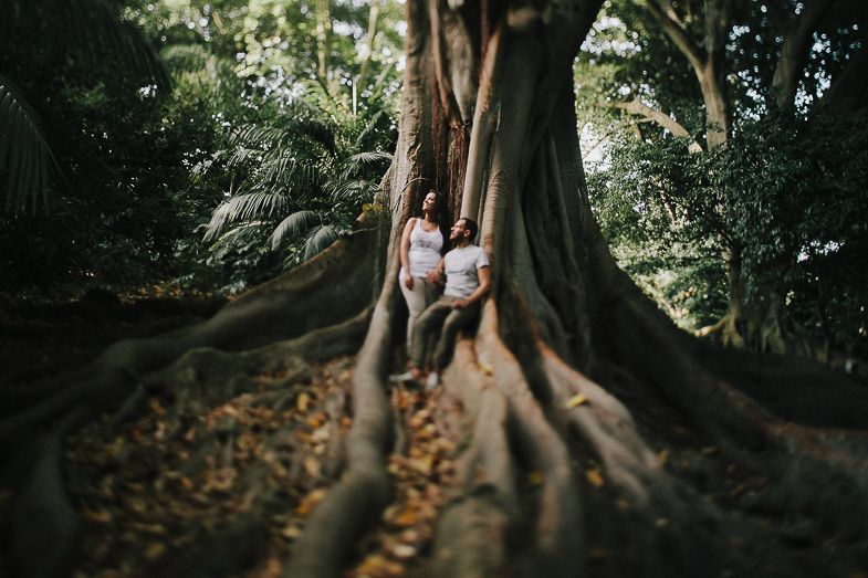 A couple poses together against the massive roots of an ancient tree in a forest setting.