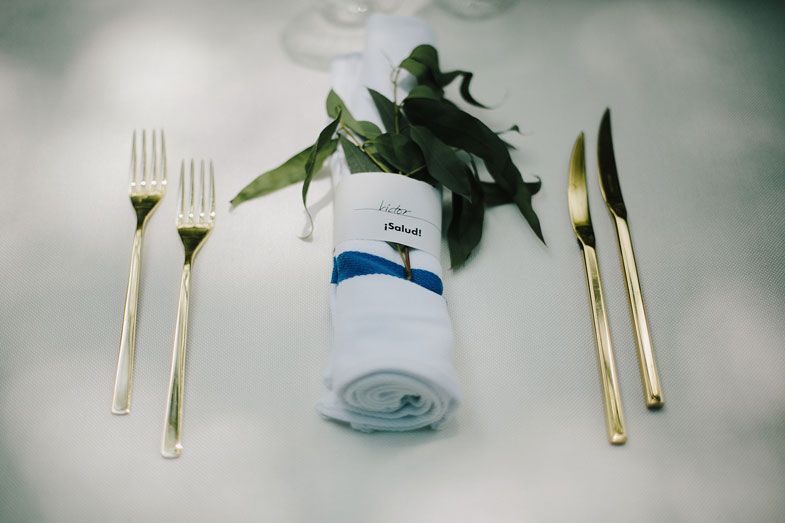 Table setting with golden cutlery and rolled white napkin with decorative green foliage.