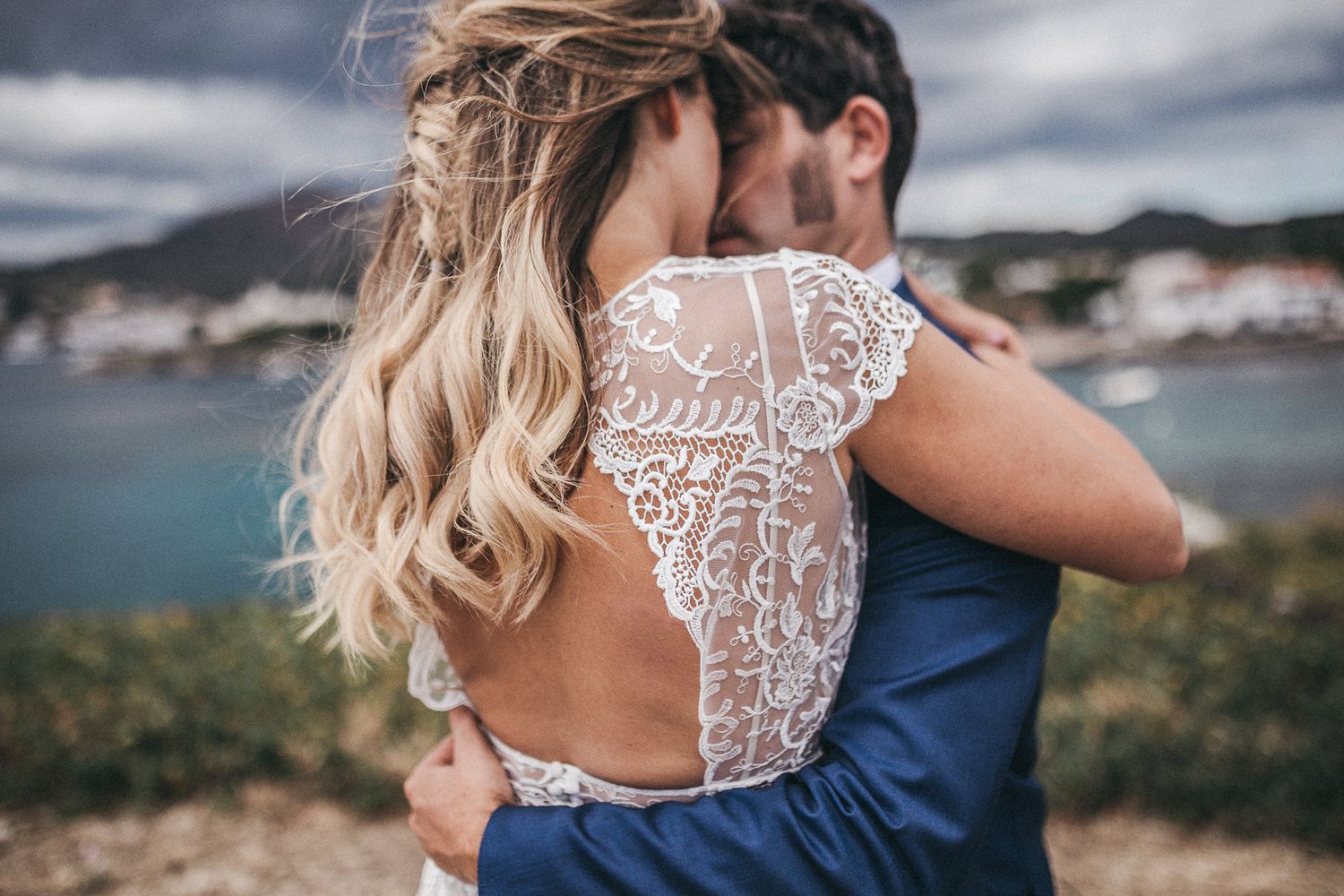 Couple embracing with scenic coastal view, bride wearing detailed lace back wedding dress.