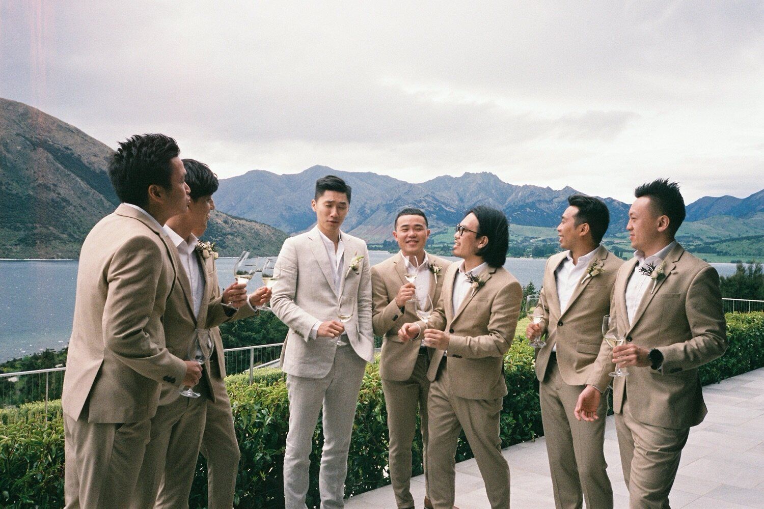 A wedding party in beige suits gathered on a scenic overlook with mountains and lake views.