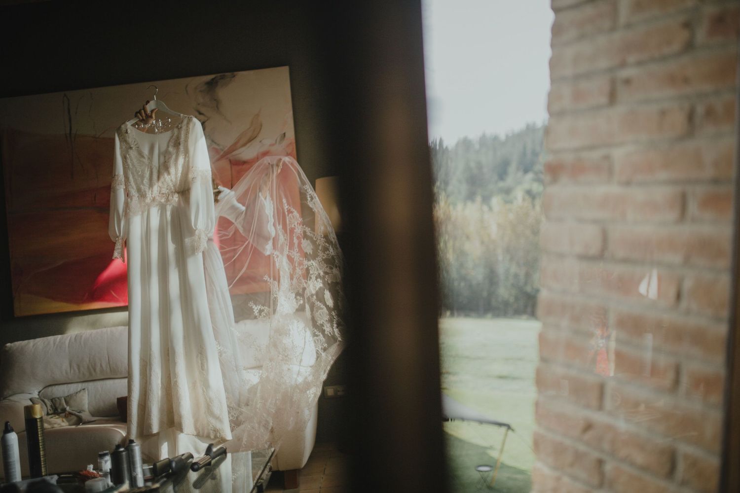 White wedding dress hanging by a window with natural light streaming in through brick walls.