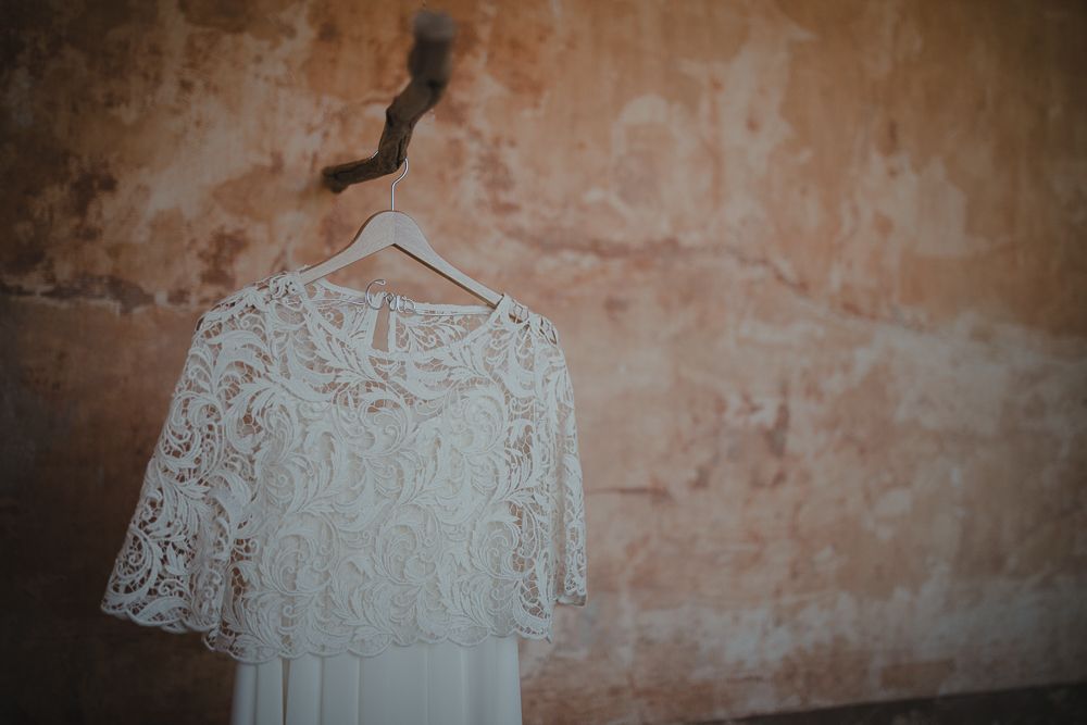 A delicate white lace wedding dress hangs against a weathered beige wall.