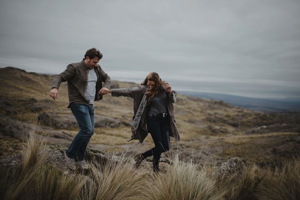 A playful couple jumps and dances through tall grass on a hillside under grey skies.