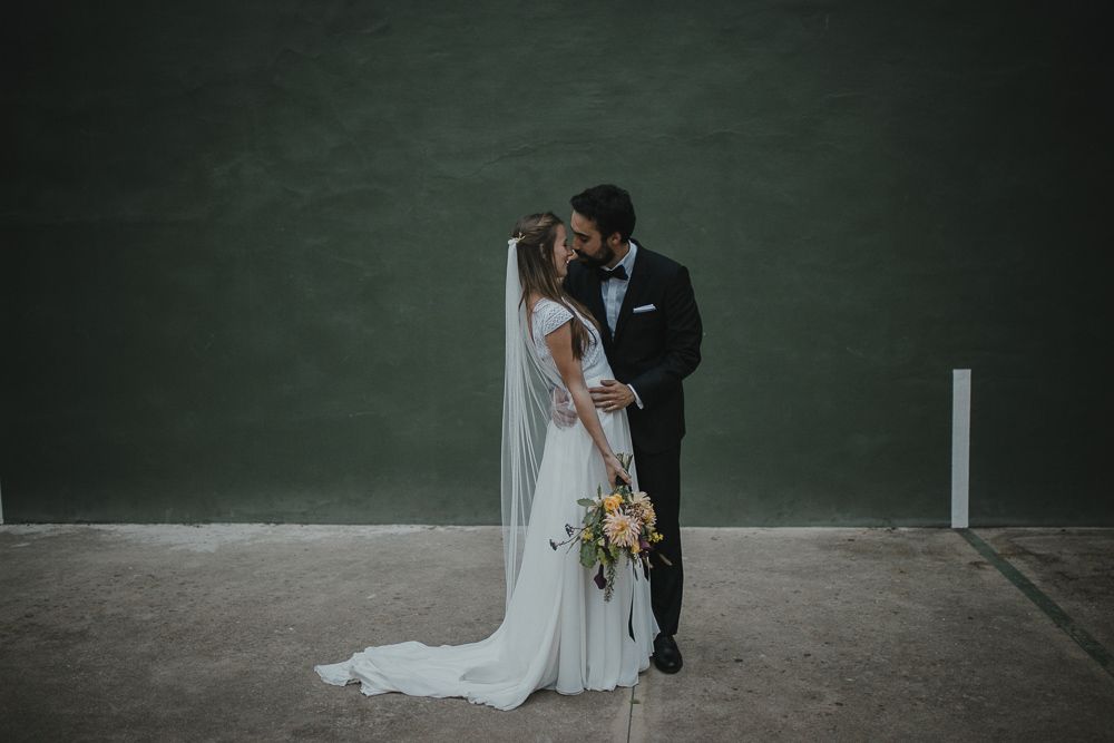 Wedding couple embracing against dark green wall in elegant white dress and black suit.
