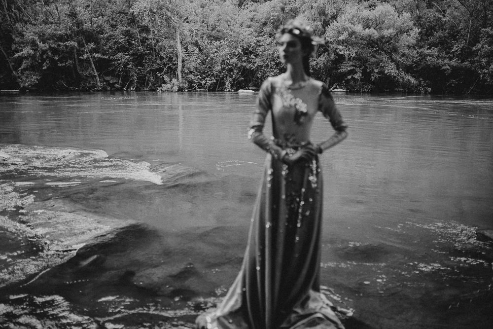Figure in flowing dress stands by water's edge in black and white photograph.