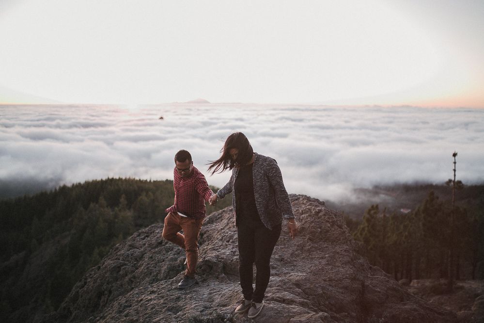 Two people walking along rocky mountain ridge above misty clouds at dawn.