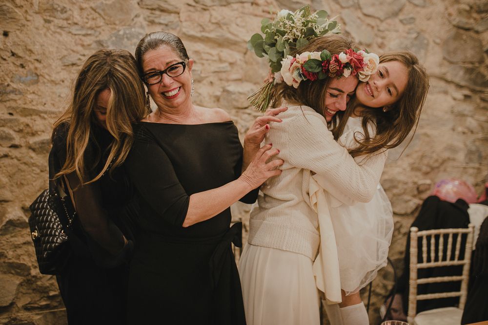 Wedding party shares laughter and hugs in front of stone wall during celebration.