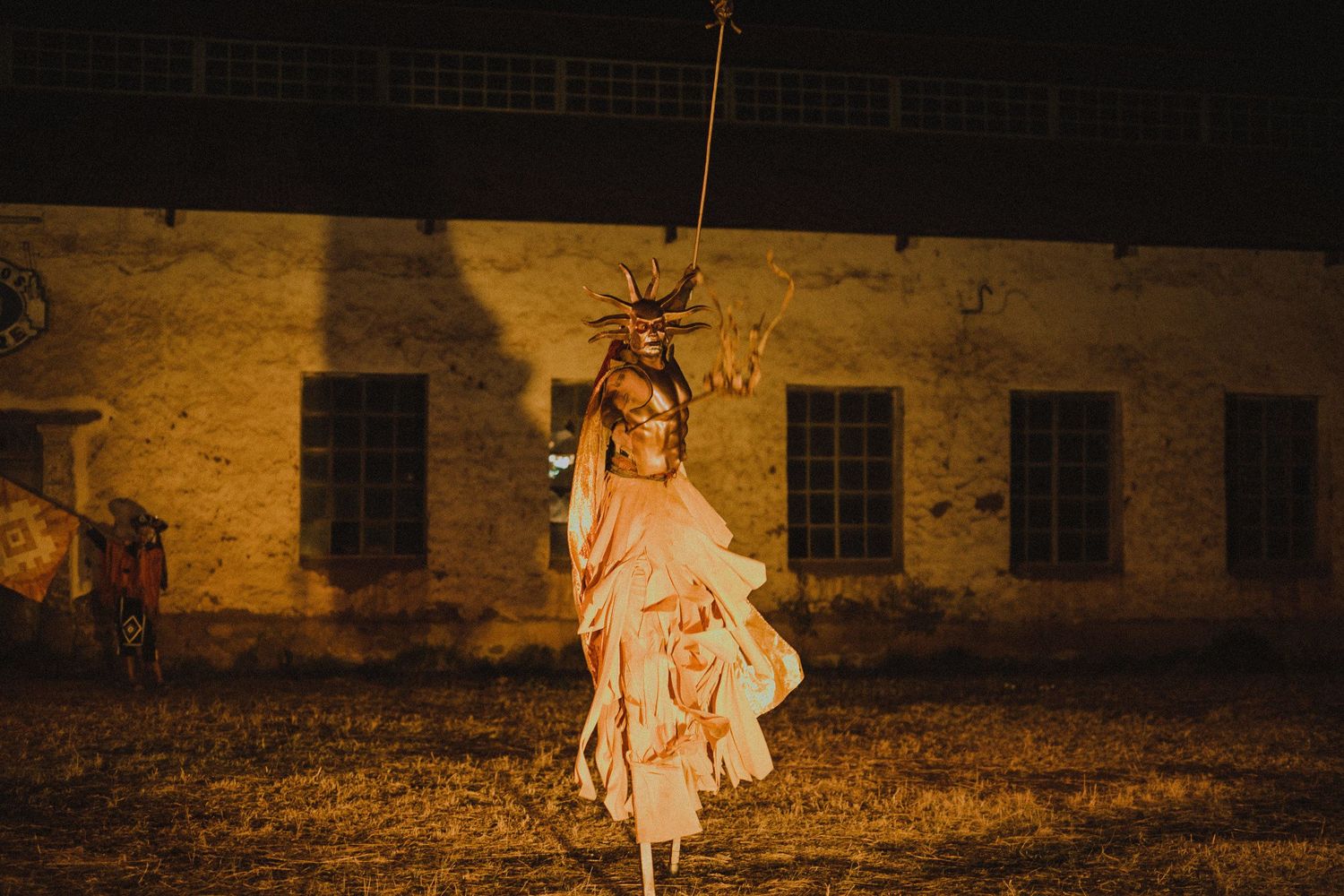 A figure wearing a flowing dress performs a dramatic suspended dance movement in front of an illuminated brick building.