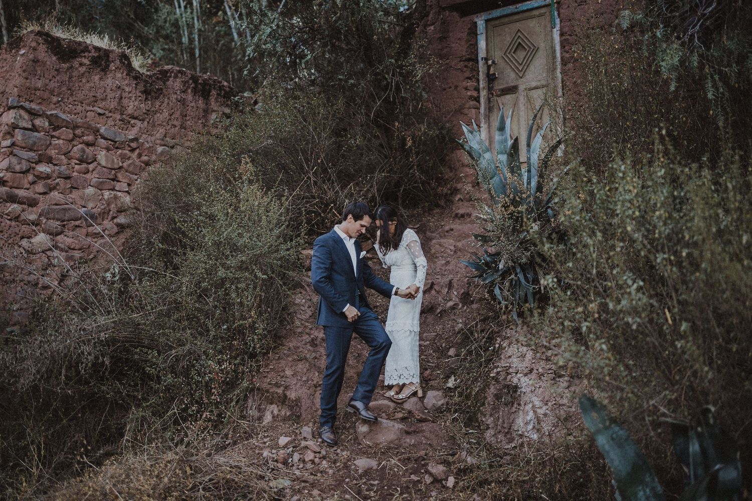 A couple in formal attire poses against a rustic brick wall surrounded by greenery.