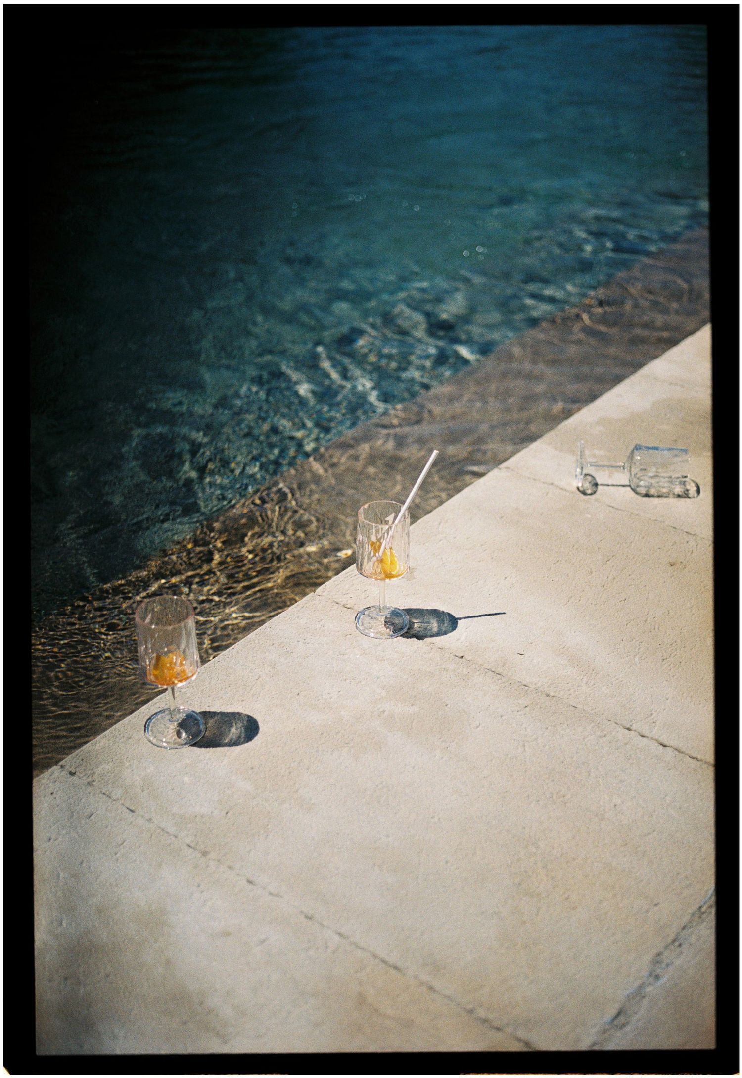 Two cocktail glasses with straws sit on a sunlit poolside ledge beside crystal clear blue water.