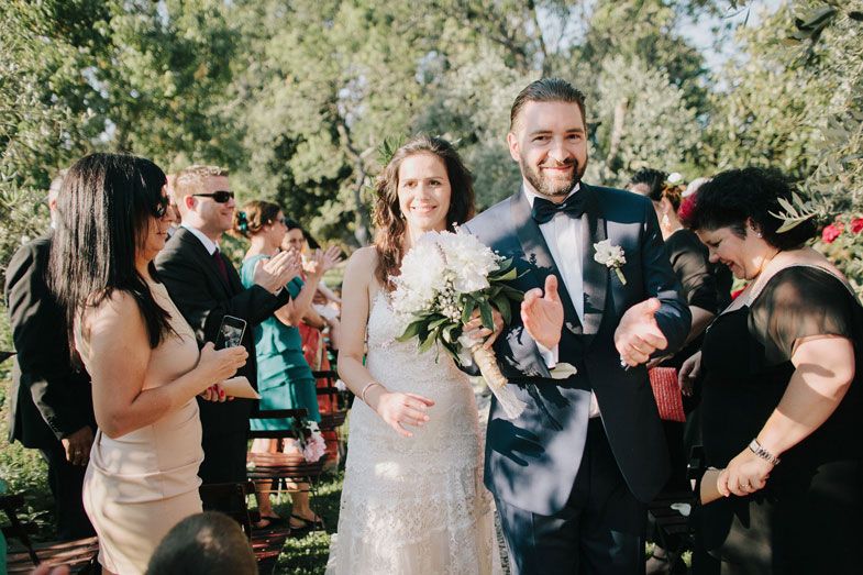 Newlyweds walk hand in hand down a garden path lined with olive trees.
