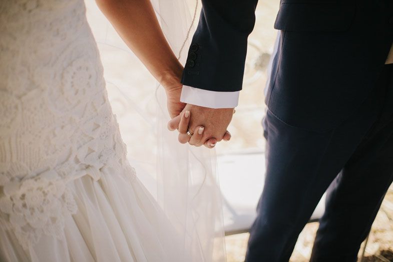 Close-up shot of newlyweds holding hands, showing white wedding dress and dark suit details.