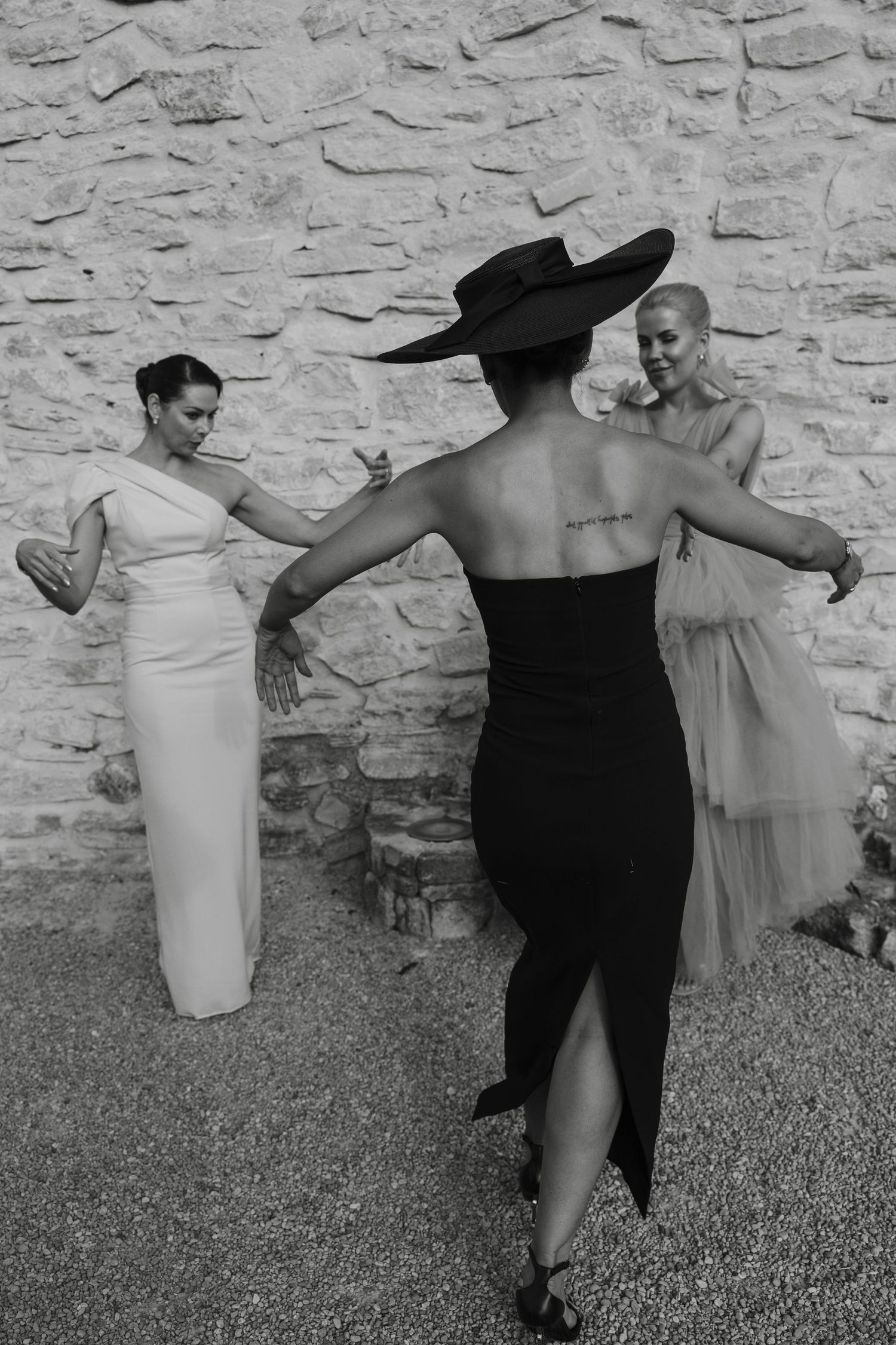 Three women in elegant dresses share a playful moment against a stone wall.