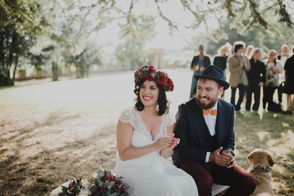 Newlywed couple sits together at an outdoor wedding ceremony with a blurred group of guests behind them.