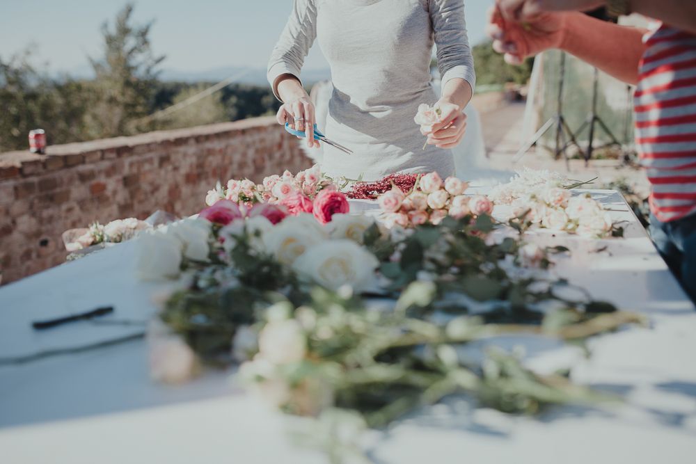 Hands arranging fresh flowers and greenery on a white table outdoors.