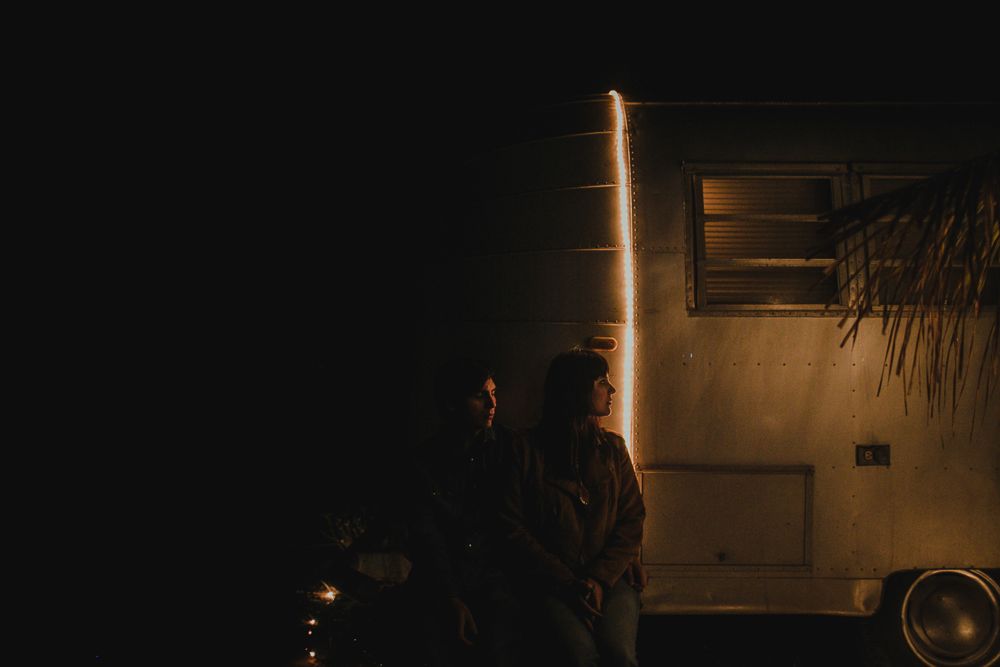 Silhouette figures stand near a vintage trailer at night with dramatic lighting effects.