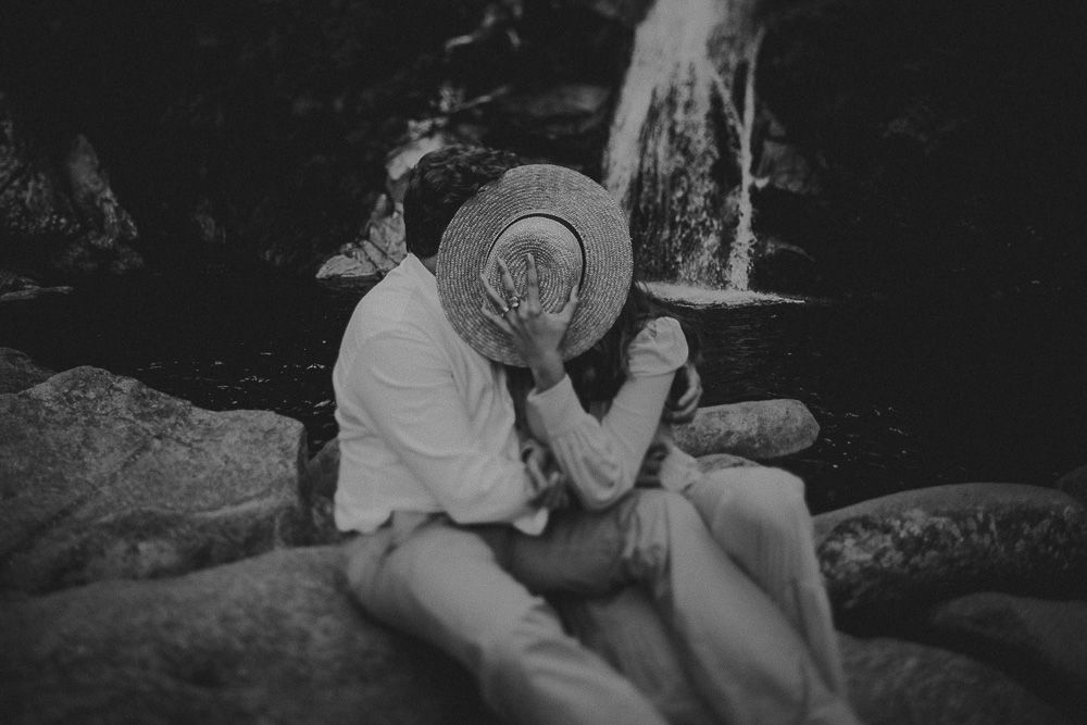 A romantic couple shares an intimate moment while sitting on rocks near a waterfall in black and white.