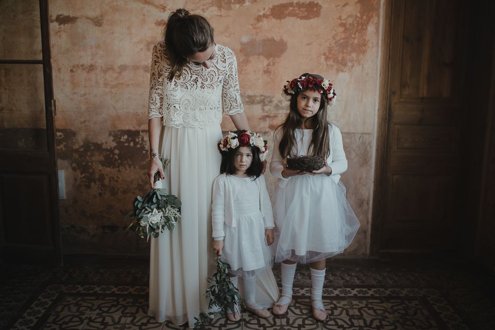 Bride in lace dress stands with two flower girls wearing floral crowns.
