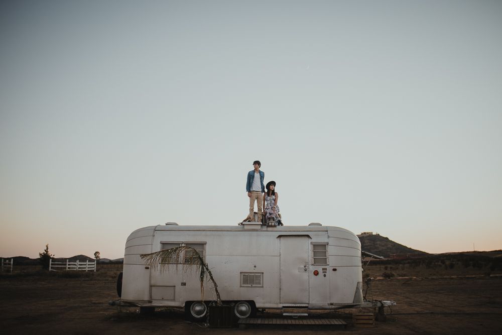 Two people stand atop a vintage Airstream trailer in a desert landscape at dusk.