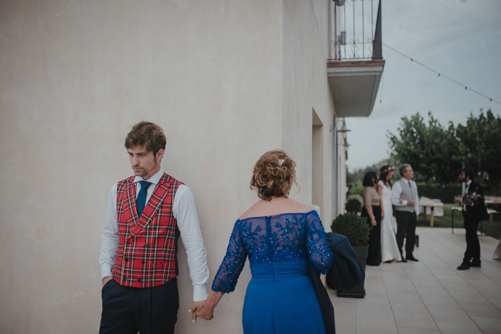 A couple holds hands while wearing red plaid vest and blue dress against a white wall.