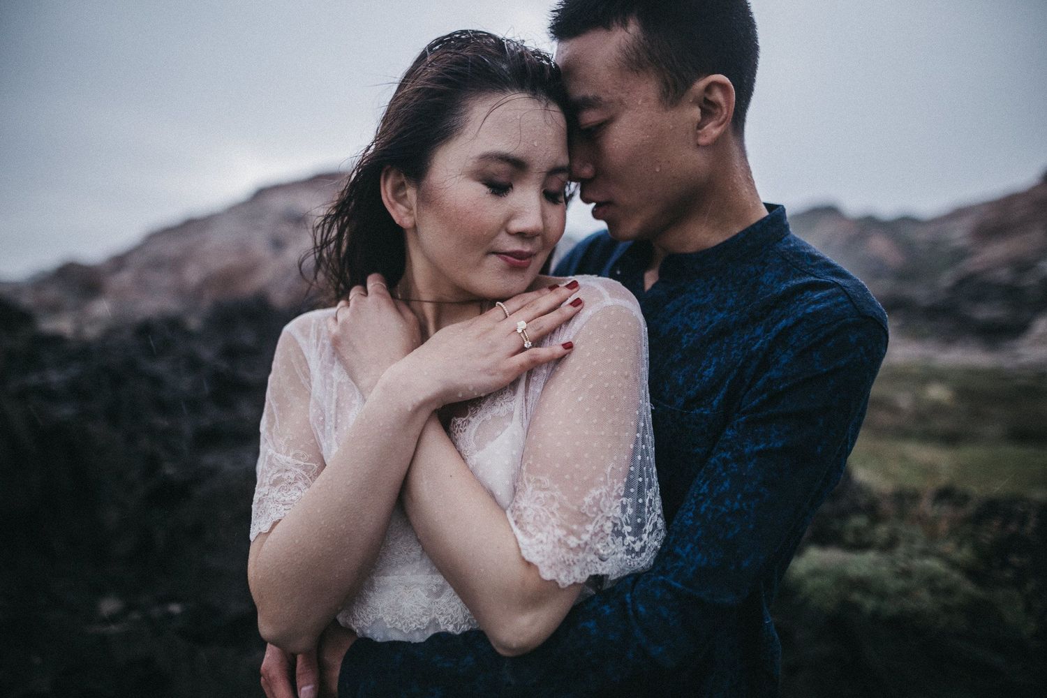 A tender moment between a couple in formal wear against a rocky landscape backdrop.